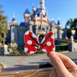 May include: A red and white polka dot fabric bow patch with black embroidered outlines. The bow features a central knot detail and two symmetrical loops, crafted with dense thread stitching throughout the entire surface of the design.