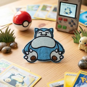 May include: A blue and cream-colored embroidered patch shaped like the character Snorlax, featuring a sleeping expression, dark blue outlines, and brown paw pads, positioned among various trading cards, a red and white ball, and a handheld device.
