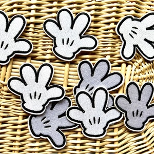 May include: A collection of embroidered patches shaped like cartoon gloves. The patches are primarily white with black outlines and details, and some are gray. They are arranged on a woven, light brown surface.