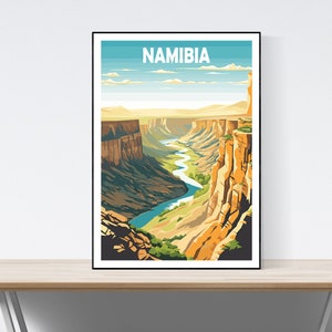 Peut inclure: Impression d'art encadrée représentant un paysage stylisé de la Namibie. L'œuvre représente un canyon avec une rivière sinueuse, sous un ciel bleu. Le mot "NAMIBIA" est imprimé en haut en lettres blanches. La palette de couleurs comprend des nuances de bleu, vert et marron.