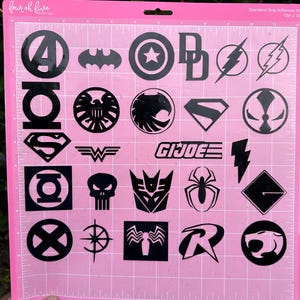 May include: A pink cutting mat featuring black superhero and comic book logos, such as the Avengers' A, Batman's bat, and Captain America's shield. The mat has a grid pattern and the text "four oh five BASICS" and "Standard Grip Adhesive Mat".