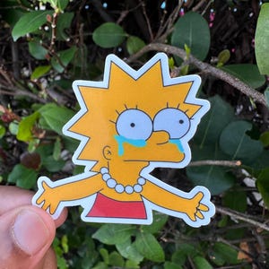 May include: A sticker featuring a cartoon character with yellow hair, large blue eyes, and tears. The character is wearing a red dress and a pearl necklace. The sticker has a white border and is set against a background of green leaves.