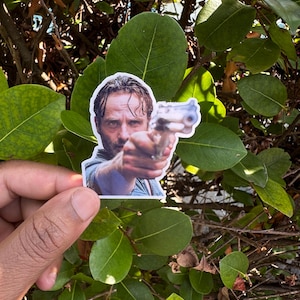 May include: A sticker featuring a man pointing a gun, with a determined expression. The sticker is held in front of a backdrop of green leaves and branches, creating a contrast between the image and the natural setting.