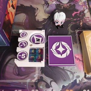May include: A purple card with a white diamond symbol in the center. The card is next to a white plastic tray with a purple diamond symbol and three purple dice with white numbers. The tray also has three white circles with the numbers -1, -3, and -5. The card and tray are on a purple and white game board with the numbers 6 through 32.