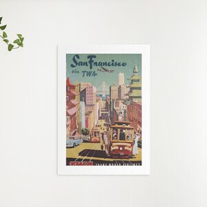 May include: Vintage travel poster advertising a trip to San Francisco via TWA. The poster features a colorful illustration of a cable car traveling up a hill with the Golden Gate Bridge in the background. The text "San Francisco via TWA" is printed in bold letters at the top of the poster.