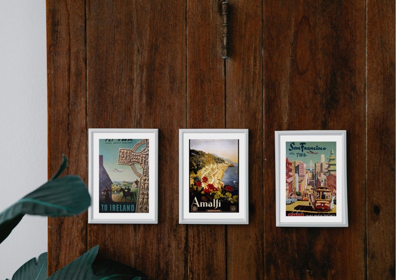 May include: Three framed vintage travel posters on a wooden wall. The posters feature images of a cross in Ireland, a coastal town in Amalfi, and a cityscape of San Francisco.