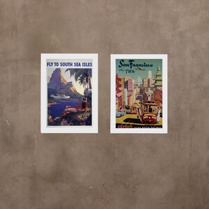 May include: Two vintage travel posters framed in white. The left poster features a tropical island scene with a plane flying overhead and the text "Fly to South Sea Isles". The right poster features a cityscape with a cable car and the text "San Francisco via TWA".