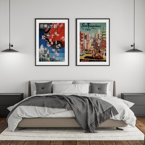 May include: Two framed vintage travel posters hanging on a white wall above a bed with a gray and white blanket. The poster on the left is in Japanese and features a red umbrella and cherry blossoms. The poster on the right is for San Francisco and features a cable car.