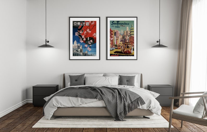 May include: Two framed vintage travel posters hanging on a white wall above a bed with a gray and white blanket. The poster on the left is in Japanese and features a red umbrella and cherry blossoms. The poster on the right is for San Francisco and features a cable car.