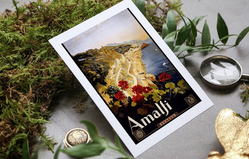 May include: A vintage travel poster featuring a scenic view of Amalfi, Italy. The poster is printed on white cardstock and features a colorful illustration of a coastal town with a winding road leading up a hillside. The text "Amalfi" is printed in bold, black letters at the bottom of the poster.