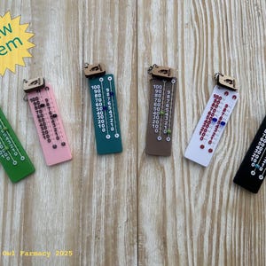 May include: A collection of colorful, miniature thermometers with wooden tops, each displaying a numbered scale from 10 to 100. The thermometers are in green, pink, teal, brown, white, and black. A yellow starburst with "New Item" is in the upper left.