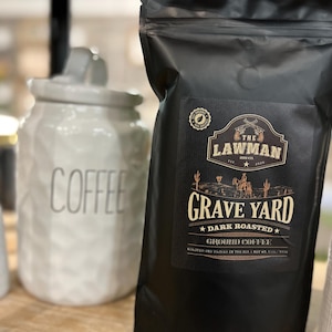 May include: A black bag of Grave Yard dark roasted ground coffee from The Lawman Coffee Co. The bag features a western-themed design with a cowboy riding a horse and cacti in the background. The text "Grave Yard" is in a large, bold font.