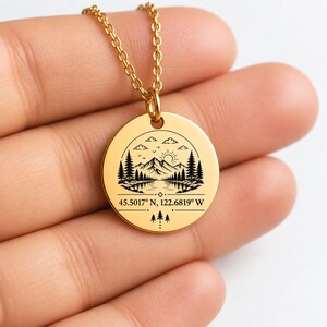Könnte beinhalten: Eine goldfarbene, runde Anhängerkette mit einer schwarzen Gravur eines Landschaftsmotivs aus Bergen, Tannen, einer Sonne und einem See. Unter der Landschaft steht der Text 45.5017° N, 122.6819° W, gefolgt von drei kleinen Tannen-Symbolen.