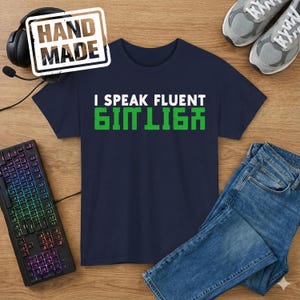 May include: Navy blue t-shirt with the text "I SPEAK FLUENT BITLISH" in white and green. The shirt is displayed with a keyboard, headphones, jeans, and sneakers on a wooden surface. A "HAND MADE" sticker is also visible.