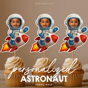 Puede incluir: Tres cupcakes con decoraciones personalizadas con temática de astronauta. Cada decoración presenta a un niño sonriente con casco y traje espacial, montado en un cohete rojo y blanco. Los cupcakes tienen un remolino de glaseado de vainilla. El texto "personalizado ASTRONAUTA" es visible.