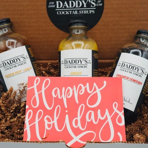 May include: A gift box with three glass bottles of Daddy's Cocktail Syrups: Passion Fruit-Vanilla, Ginger, and Cinnamon Demerara. A coral card with white script reading "Happy Holidays" is in front of the bottles. The box is filled with brown crinkle paper.