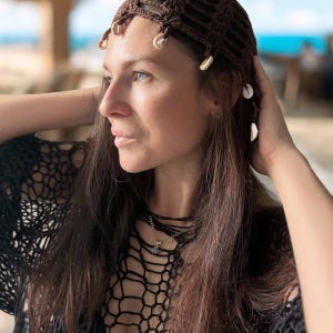 May include: A dark brown crochet headpiece featuring a grid-like pattern and small white cowrie shells attached along the front edge. The headpiece is worn over long brown hair, paired with a matching dark brown crochet top with a wide net design.