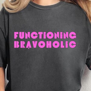 Puede incluir: Camiseta gris oscuro con el texto "FUNCTIONING BRAVOHOLIC" en letras rosa brillante. La camiseta es de manga corta y cuello redondo. La persona lleva vaqueros azules.