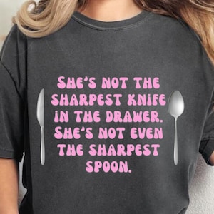 Puede incluir: Una camiseta gris oscuro con un gráfico rosa que dice "She's not the sharpest knife in the drawer. She's not even the sharpest spoon." El gráfico está acompañado por un tenedor y una cuchara plateados.