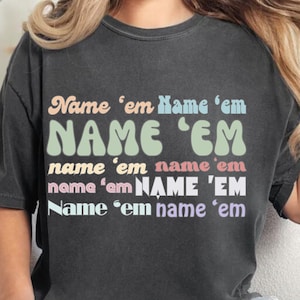 Puede incluir: Una camiseta gris oscuro con el texto "Name 'em Name 'em NAME 'EM name 'em name 'em name 'am NAME 'EM Name 'em name 'em" impreso en varios colores pastel.