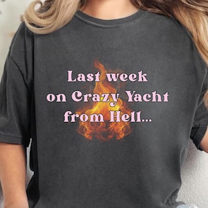Op de afbeelding: Een zwart t-shirt met een verweerde look met een roze afbeelding van een vlam met de tekst "Last week on Crazy Yacht from Hell..."