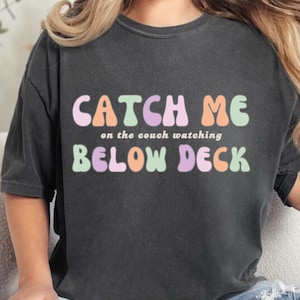 Może przedstawiać: Czarny T-shirt z napisem "Catch Me on the couch watching Below Deck" w kolorowych, bąbelkowych literach.