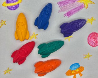 May include: Six colorful rocket-shaped crayons on a white background with crayon drawings of stars, a planet, and a comet.