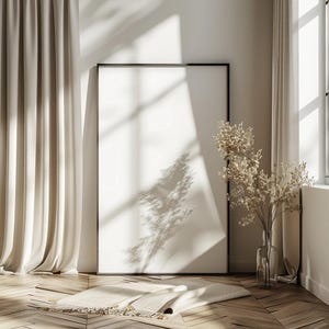 May include: A large, black framed picture hangs on a white wall in a room with beige curtains and a vase of dried flowers. The picture is blank and the room is lit by sunlight streaming through a window.