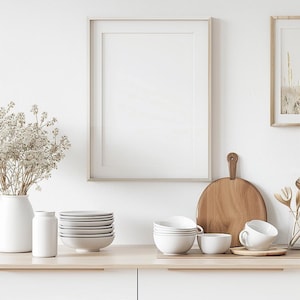 May include: A white kitchen counter with a framed picture, a wooden cutting board, a vase with white flowers, and a stack of white bowls. There are also two white mugs and a white ceramic jar on the counter.