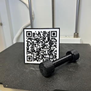May include: A black and white QR code on a white square with a black border, next to a small black dumbbell. The objects are on a dark gray surface, likely a 3D printer bed. The dumbbell has textured handles.