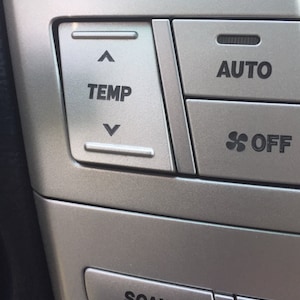 May include: Close-up of a car's climate control panel with buttons for temperature, auto, and fan speed. The buttons are silver and have black lettering. The text on the buttons reads "TEMP", "AUTO", and "OFF".
