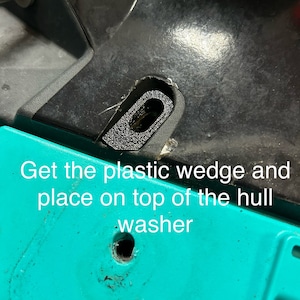 May include: A close-up of a black plastic wedge placed on top of a turquoise surface. The text 'Get the plastic wedge and place on top of the hull washer' is visible below the wedge.