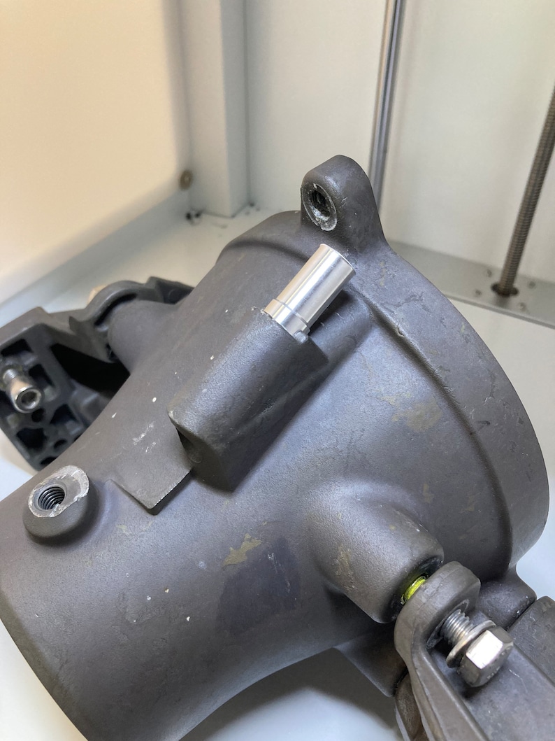 Puede incluir: Primer plano de una pieza de metal gris con una varilla de metal plateado insertada en un agujero. La pieza tiene una forma redondeada y parece ser parte de una m&aacute;quina m&aacute;s grande.