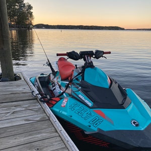 May include: A turquoise and black Sea-Doo Spark watercraft with red and white accents. The watercraft has the text 'Sea-Doo' and 'Spark' on the side and 'SC 2677 DG' on the front. The watercraft is docked at a wooden pier.