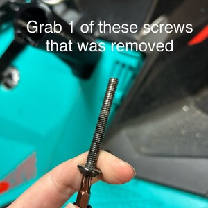 May include: A black screw with a silver head and a silver Phillips head screwdriver bit attached to it. The screw is held in a hand against a turquoise background.