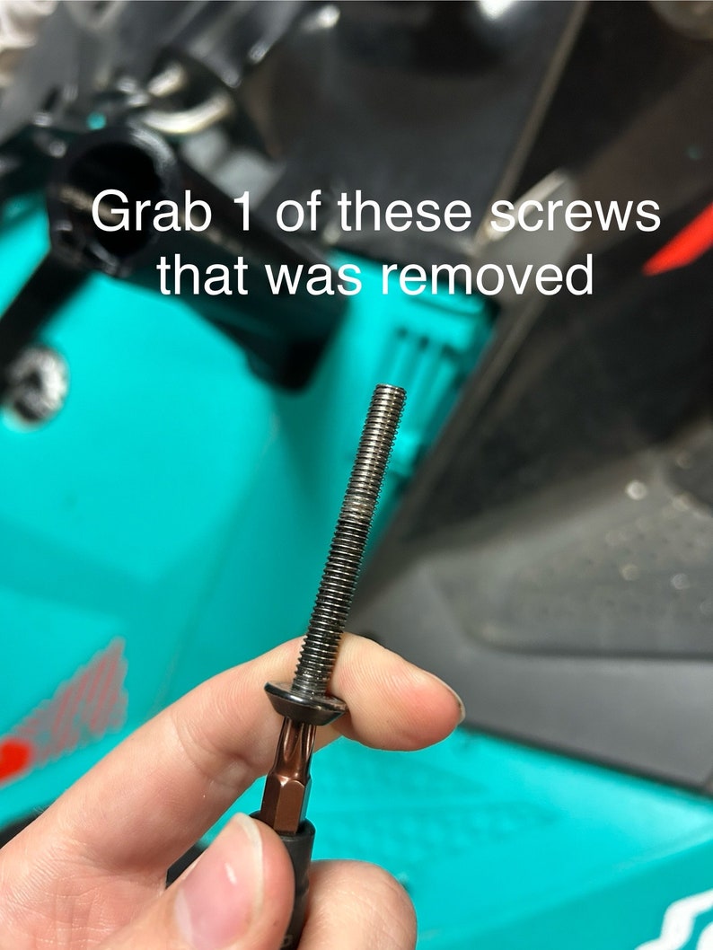 May include: A black screw with a silver head and a silver Phillips head screwdriver bit attached to it. The screw is held in a hand against a turquoise background.