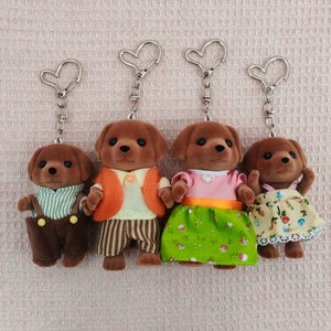 May include: Four brown plush dog keychains with different outfits. The dogs are wearing a striped shirt with overalls, an orange waistcoat with striped trousers, a pink shirt with a green skirt, and a yellow dress with floral print.