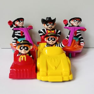Op de afbeelding: Een verzameling van vijf plastic McDonald's Hamburglar-figuurtjes. Drie figuren rijden op paarse en oranje steppen, terwijl twee figuren in kleine rode en gele auto's zitten. Elk personage draagt een zwart masker, een hoed en een gestreepte outfit.