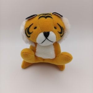 Könnte beinhalten: Ein kleines Plüsch-Tiger-Spielzeug mit orangefarbenem Fell, schwarzen Streifen auf Stirn und Wangen, einer weißen Schnauze, einer weißen Brust und weißen Fellbüscheln an den Kopfseiten. Der Tiger hat eine schwarz gestickte Nase und einen Mund.