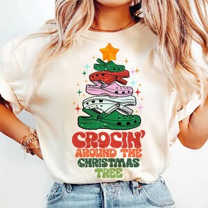 Peut inclure: T-shirt crème avec un motif de sapin de Noël composé de chaussures Crocs colorées. Le motif comprend une étoile dorée et le texte "CROCIN' AROUND THE CHRISTMAS TREE" en rouge, vert et blanc.