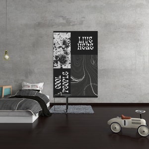 Op de afbeelding: Een zwart-witte poster met de tekst "LIVE HERE" en "COOL PEOPLS" hangt aan een grijze betonnen muur. Een wit bed met een grijs-witte bedsprei met patroon staat in de kamer. Een speelgoedauto staat op de vloer.