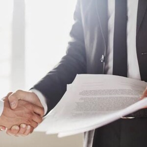 May include: Two people shaking hands in a business setting. One person is wearing a white shirt, and the other is wearing a black suit with a white shirt and black tie. The person in the suit is holding a stack of papers.