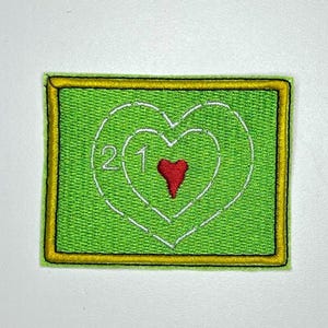 May include: Embroidered patch with a green background and a yellow border. The patch features a white heart design with a red heart in the center. The numbers "2" and "1" are inside the heart design.