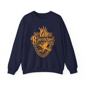 May include: Navy blue sweatshirt featuring the Ravenclaw crest in gold. The crest includes an eagle, banner, and the word "Ravenclaw". The sweatshirt is a crew neck style and has long sleeves.