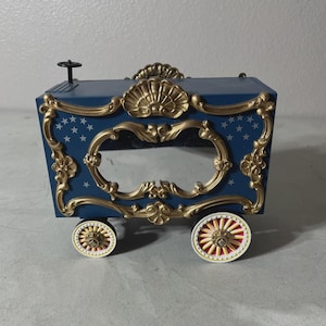 May include: A navy blue decorative music box shaped like a circus wagon. The box features gold filigree, a shell-shaped top, and star accents. The wheels are cream-colored with red and gold details.