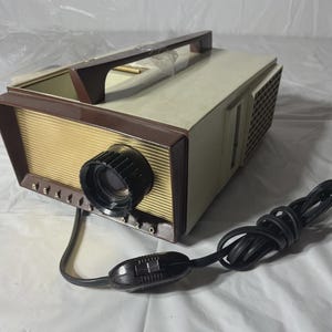 May include: Vintage film projector with a cream and brown exterior. The projector has a black lens, a handle, and a power cord with a switch. The front of the projector has a gold-colored, ribbed panel. A classic piece of home entertainment equipment.