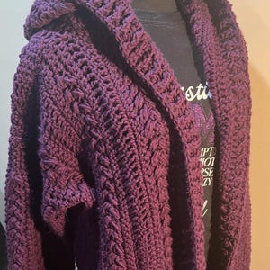 Może przedstawiać: Głęboki fioletowy kardigan z kapturem wykonany na szydełku. Ręcznie robiony sweter ma teksturowany wzór ściegu i drapowany, otwarty przód. Kaptur i rękawy są widoczne, prezentując skomplikowany wzór szydełkowy. Kardigan jest prezentowany na manekinie.