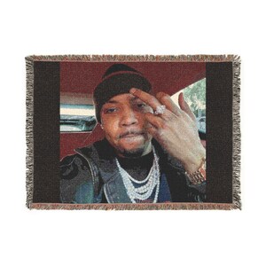 May include: Woven throw blanket featuring a portrait of a person wearing a beanie, jewelry, and a jacket. The image has a red and black color scheme. The blanket has a fringed edge.