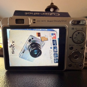 May include: A silver Sony Cyber-shot DSC-W170 digital camera with a Super SteadyShot feature. The LCD screen displays a live view of the camera's own product packaging, showing the model name, 10.1 megapixels, 5x optical zoom, and 28mm lens.