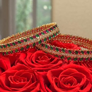 May include: Two gold-toned bangles featuring alternating marquise-cut red and green stones arranged in a leaf-like pattern, with a central row of small clear white stones running horizontally through the middle of each circular bracelet.